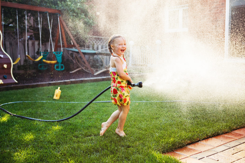 One girl splashing with gardening house on backyard on summer day. Child playing with water outside at sunset. Candid moment lifestyle home kid activity.